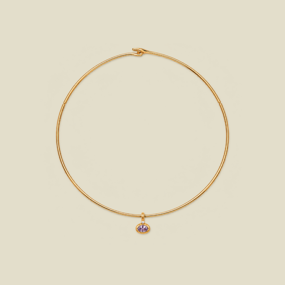 A delicate gold wire necklace with a small dangling charm featuring a light purple oval gemstone, set against a solid beige background.