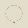 A delicate gold wire necklace with a small dangling charm featuring a light purple oval gemstone, set against a solid beige background.