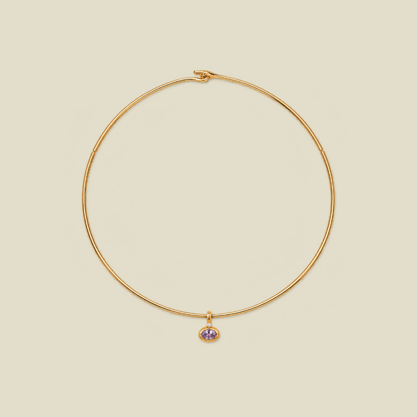 A delicate gold wire necklace with a small dangling charm featuring a light purple oval gemstone, set against a solid beige background.