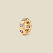 A gold ring charm with multiple bezel set square-cut light purple gemstones set side by side to create the band, shown on a solid beige background.
