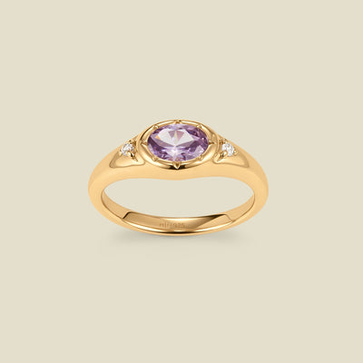 A gold signet ring with a light purple center oval bezel-set gemstone, and two small clear accent stones on either side, set against a solid beige background.