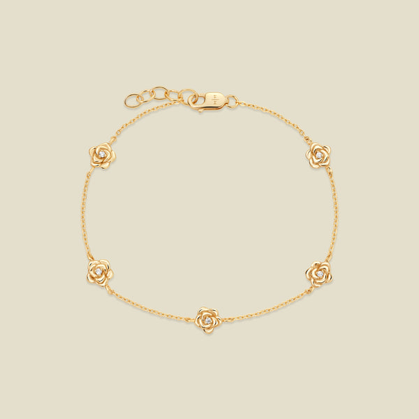A delicate gold chain bracelet featuring five ornate, textured gold rose charms with small clear stones, arranged in a circle on a light beige background.