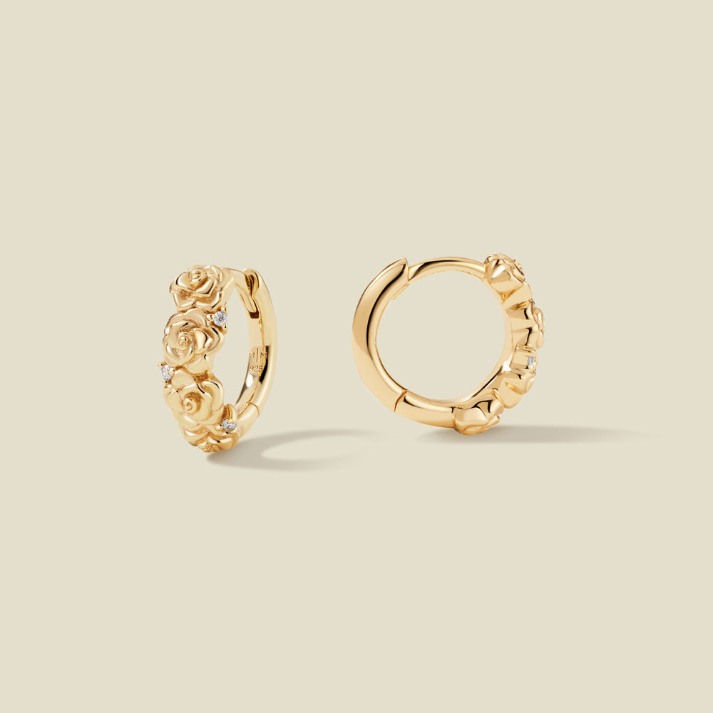 A pair of gold hoop earrings adorned with textured rose flowers stacked along the hoop, with scattered small clear accent stones, set against a solid beige background.