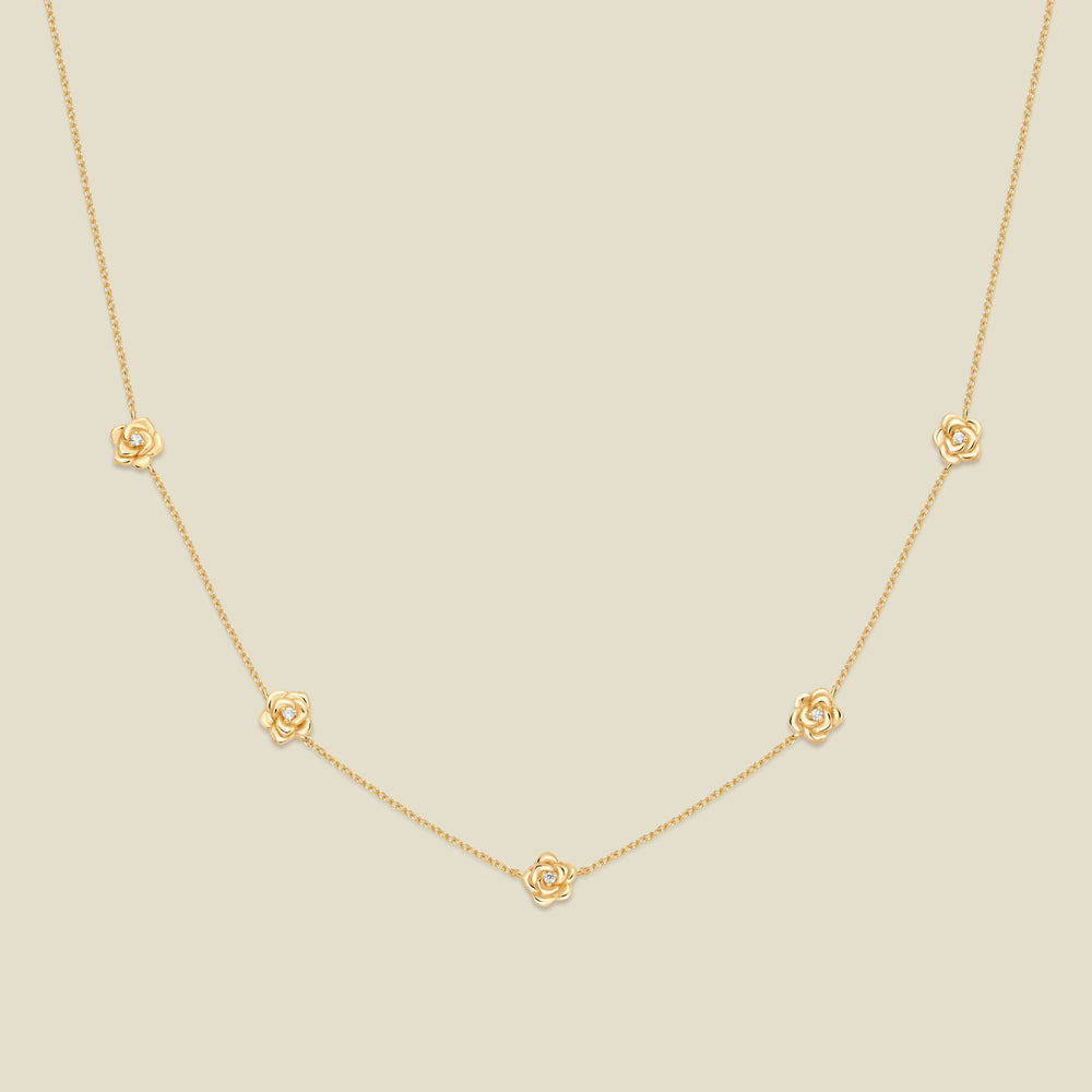 A delicate gold chain necklace featuring five small, evenly spaced textured rose flower motifs set with tiny clear stones, displayed on a solid light beige background.