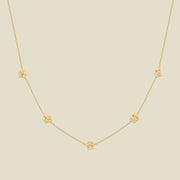 A delicate gold chain necklace featuring five small, evenly spaced textured rose flower motifs set with tiny clear stones, displayed on a solid light beige background.