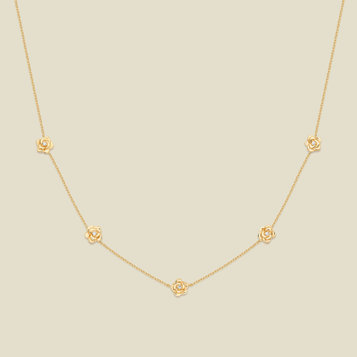 A delicate gold chain necklace featuring five small, evenly spaced textured rose flower motifs set with tiny clear stones, displayed on a solid light beige background.