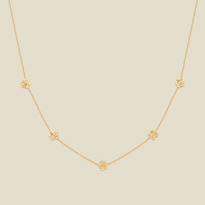 A delicate gold chain necklace featuring five small, evenly spaced textured rose flower motifs set with tiny clear stones, displayed on a solid light beige background.