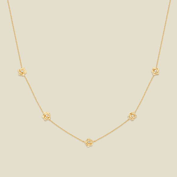 A delicate gold chain necklace featuring five small, evenly spaced textured rose flower motifs set with tiny clear stones, displayed on a solid light beige background.