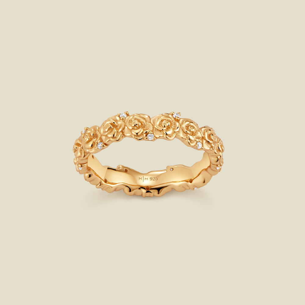 A gold floral-etched ring band with textured rose flowers and small clear gemstones, shown on a plain warm beige background. 