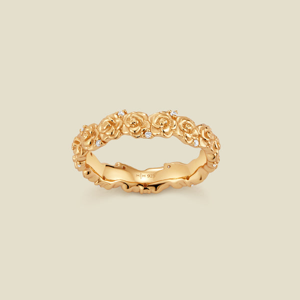 A gold floral-etched ring band with textured rose flowers and small clear gemstones, shown on a plain warm beige background. 