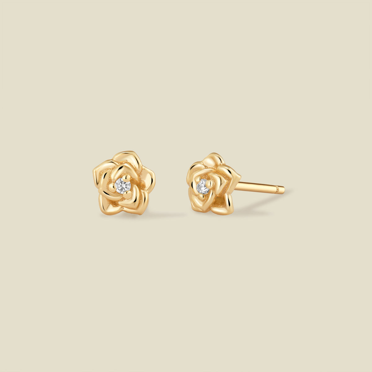 A pair of gold rose stud earrings with a small clear gemstone in the center of each rose, shown on a solid beige background.