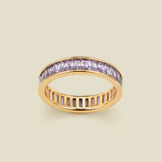 A gold eternity ring featuring a continuous channel setting of baguette-cut light purple gemstones, shown on a solid light beige background.