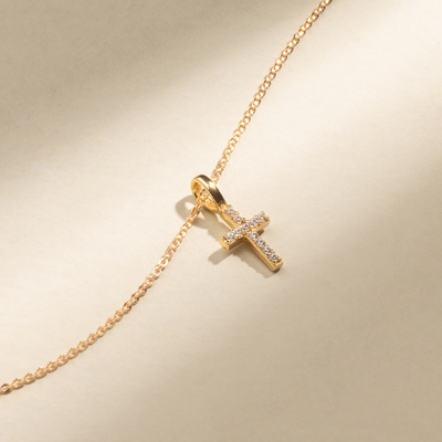 A gold chain necklace with a small pave-gemstone cross charm, set against a beige background.