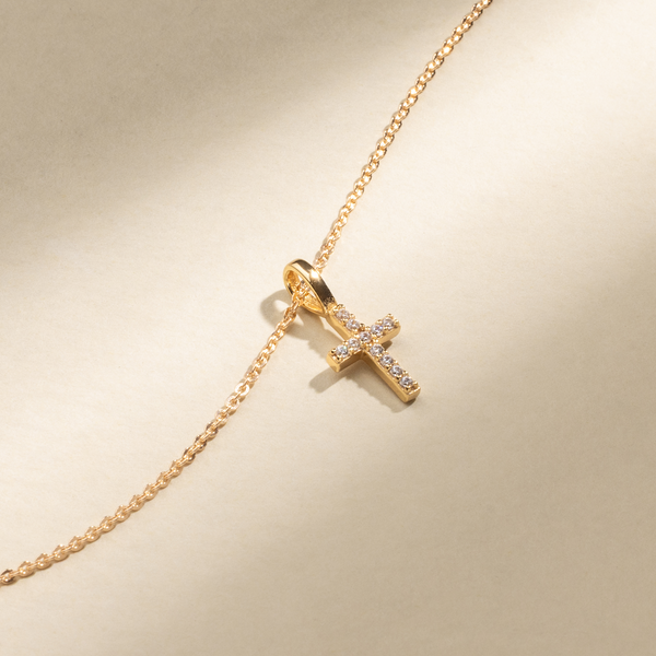 A gold chain necklace with a small pave-gemstone cross charm, set against a beige background.