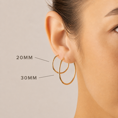 Close-up of a woman's ear wearing two gold hoop earrings, with size labels pointing to the 20MM and 30MM hoops against a soft beige background.