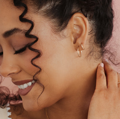 A close-up lifestyle shot of a woman wearing a delicate gold hoop earrings, showing the jewelry on her ear against a soft pink background.