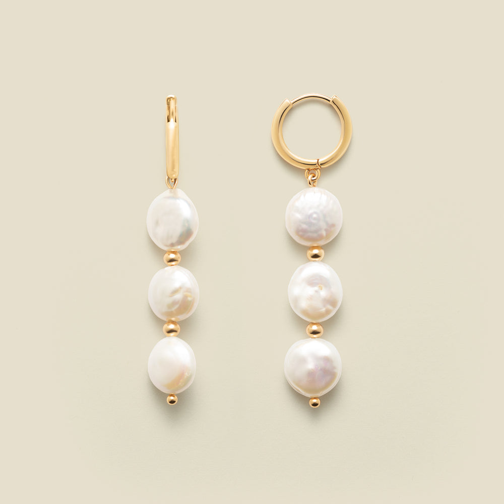 Gold hoop earrings with three dangling pearls against a neutral background.