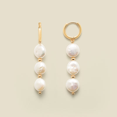 Gold hoop earrings with three dangling pearls against a neutral background.