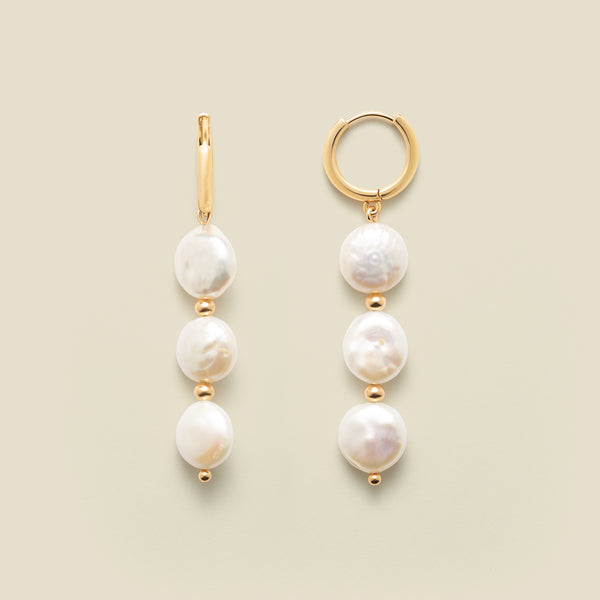 Gold hoop earrings with three dangling pearls against a neutral background.