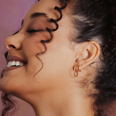 Close-up side profile of a smiling woman with closed eyes wearing two pairs of small gold hoop earrings, against a soft pink background.