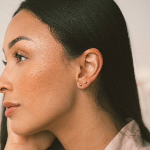 Close-up side profile of a woman wearing small gold stud earrings on her earlobe against a soft light background.