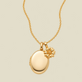 A gold necklace featuring an oval locket pendant with a small daisy charm, displayed on a light beige background.
