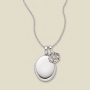 A silver locket necklace with an oval locket pendant and a small poppy flower charm, displayed on a light beige background.