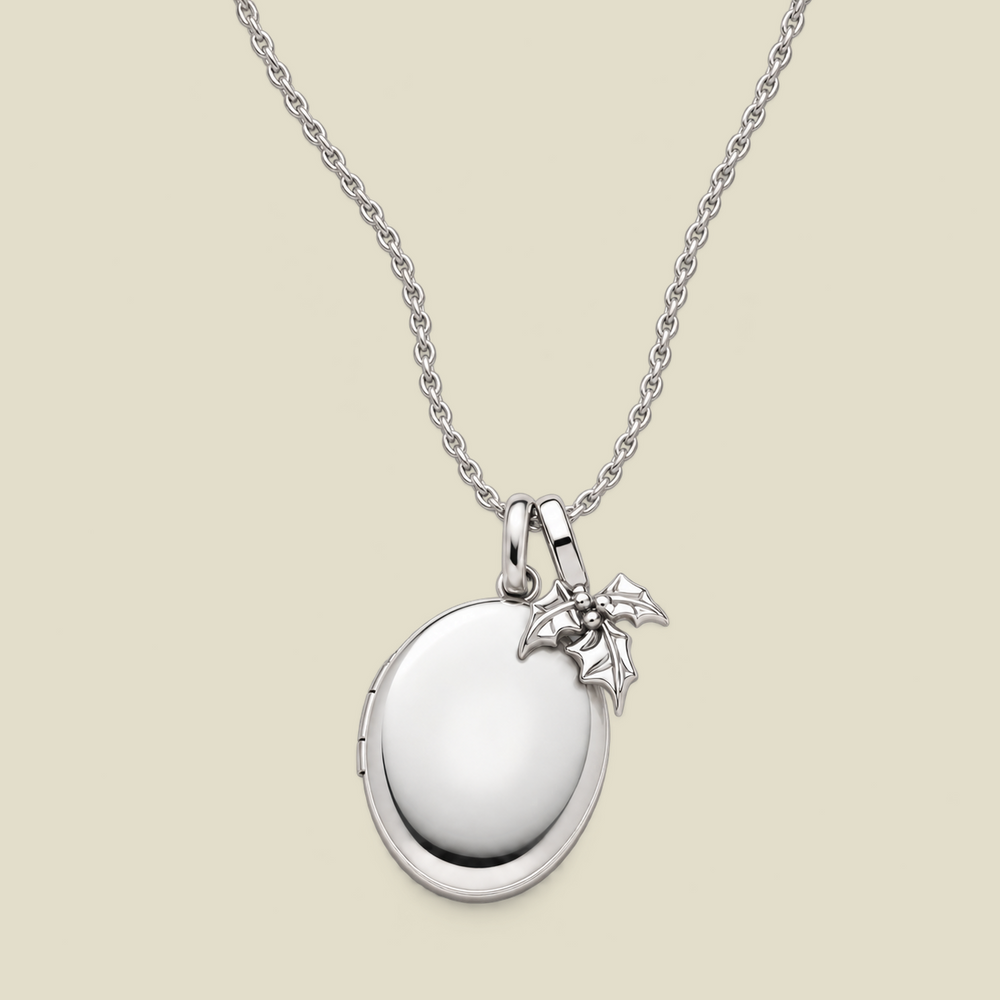 A silver locket necklace with a polished oval pendant and a small holly leaf charm, displayed on a light beige background.