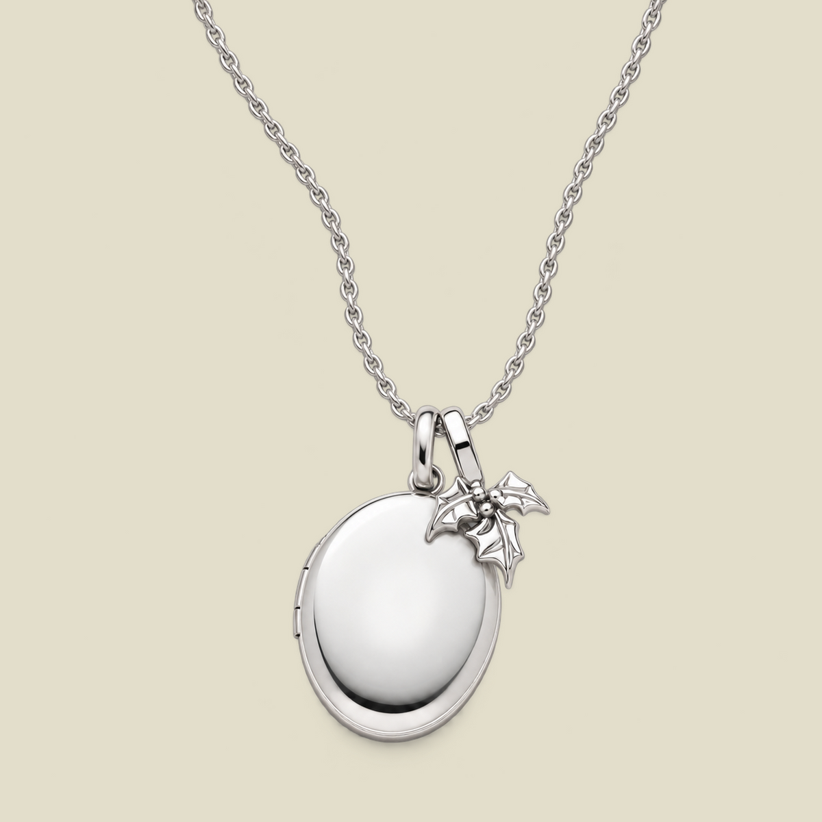 A silver locket necklace with a polished oval pendant and a small holly leaf charm, displayed on a light beige background.