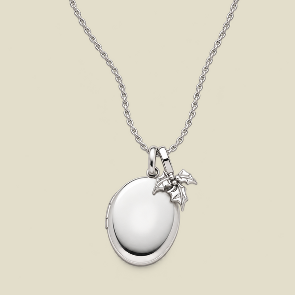 A silver locket necklace with a polished oval pendant and a small holly leaf charm, displayed on a light beige background.