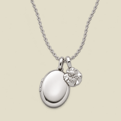 A silver locket necklace with an oval locket and a small carnation flower charm, displayed on a light beige background.