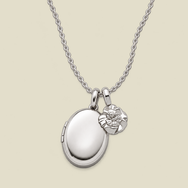 A silver locket necklace with an oval locket and a small carnation flower charm, displayed on a light beige background.