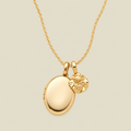 A polished gold locket necklace with an oval locket and a small carnation flower charm, displayed on a light beige background.