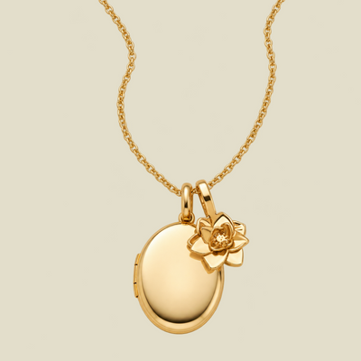 A gold locket necklace with an oval pendant and a small lotus flower charm, shown on a plain light beige background.