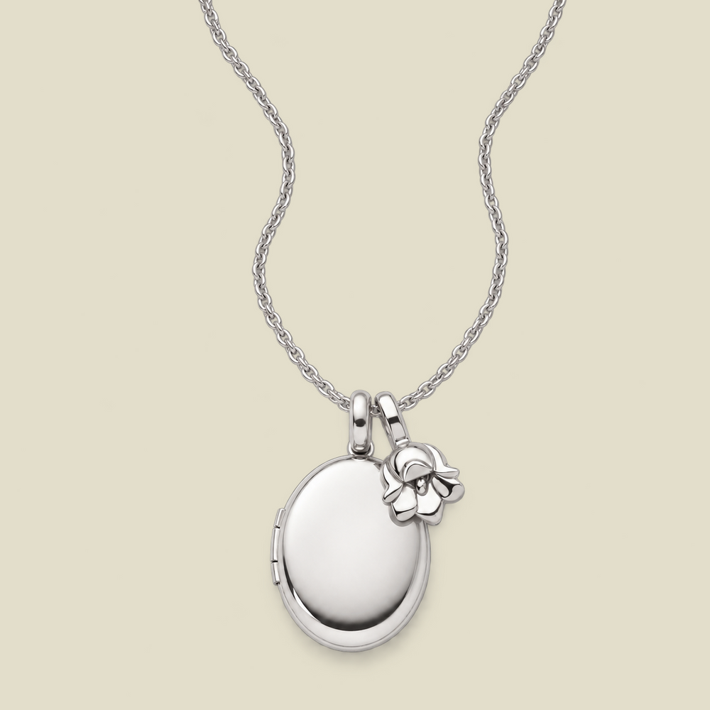A silver locket necklace with an oval locket pendant and a small lily of the valley flower charm, displayed on a light beige background.