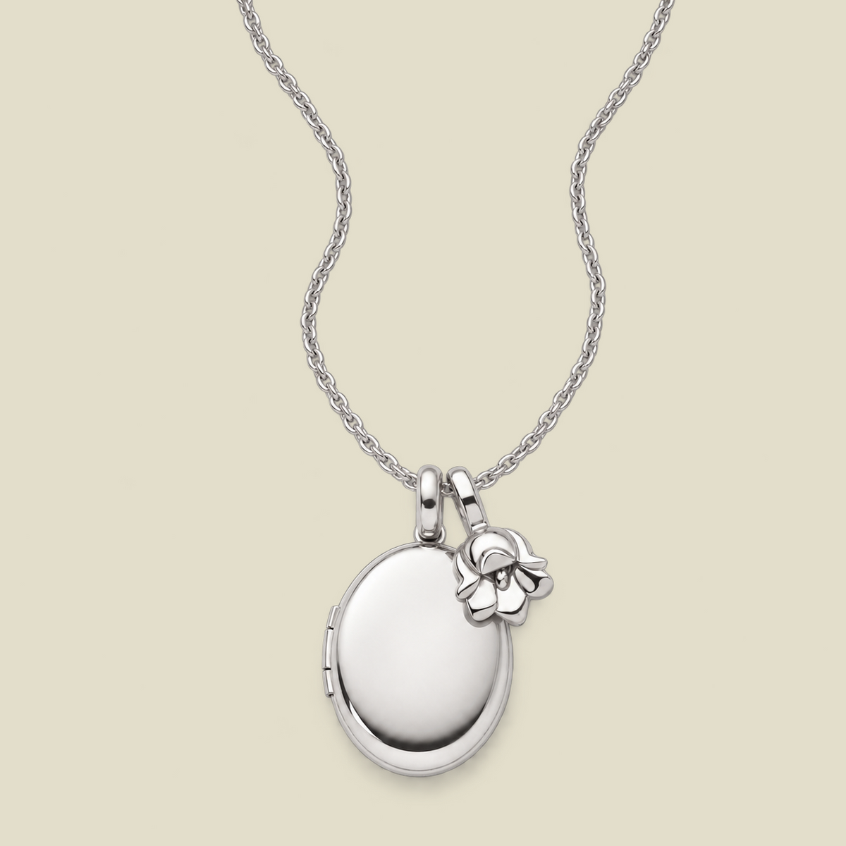 A silver locket necklace with an oval locket pendant and a small lily of the valley flower charm, displayed on a light beige background.