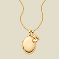 A polished gold locket necklace with a small lily of the valley flower charm, displayed on a light beige background.