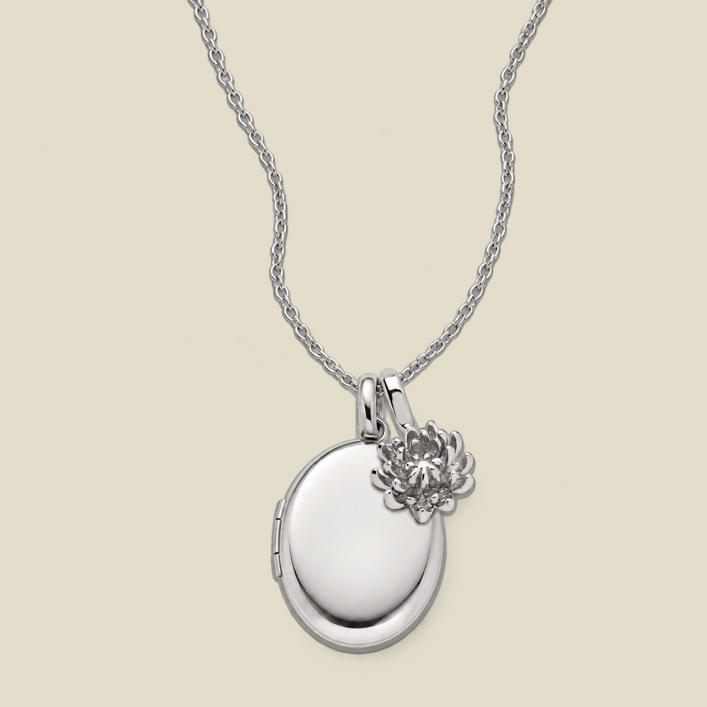 A silver oval locket necklace with a small chrysanthemum flower charm, displayed on a light beige background.
