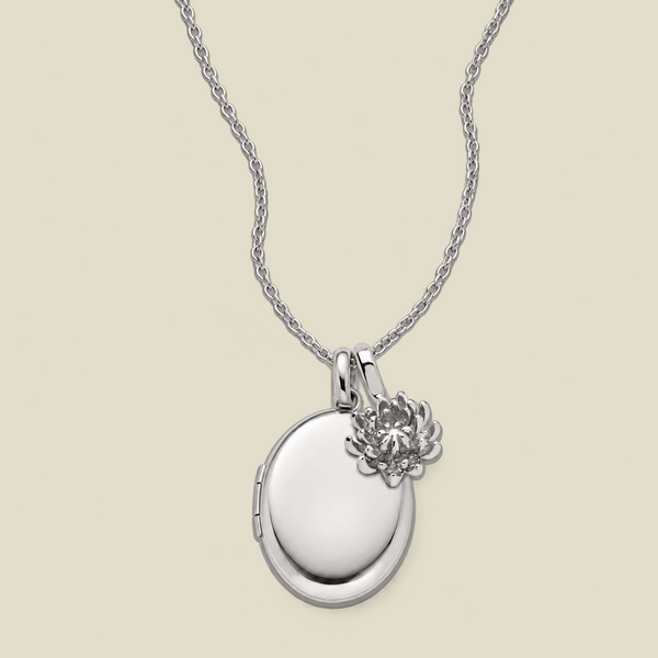 A silver oval locket necklace with a small chrysanthemum flower charm, displayed on a light beige background.
