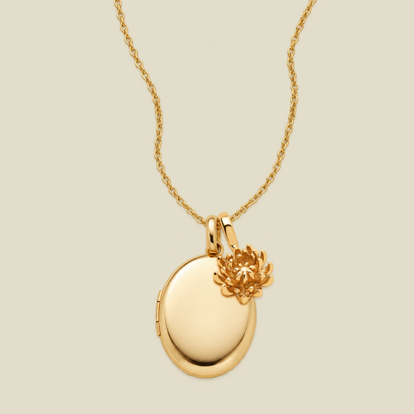 A gold locket necklace with an oval pendant and a small chrysanthemum flower charm, shown on a light beige background.
