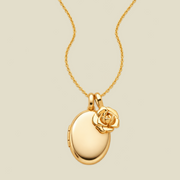 A gold oval locket necklace with a small peony flower charm, shown on a light beige background.