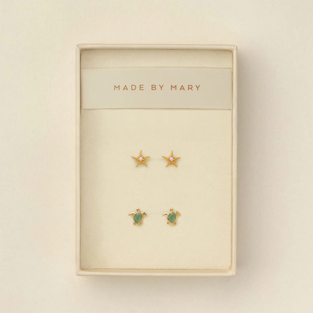 Set of gold stud earrings, one a starfish with a pearl in the center and the other a turtle with a green stone, in a 'Made by Mary' box on a beige background