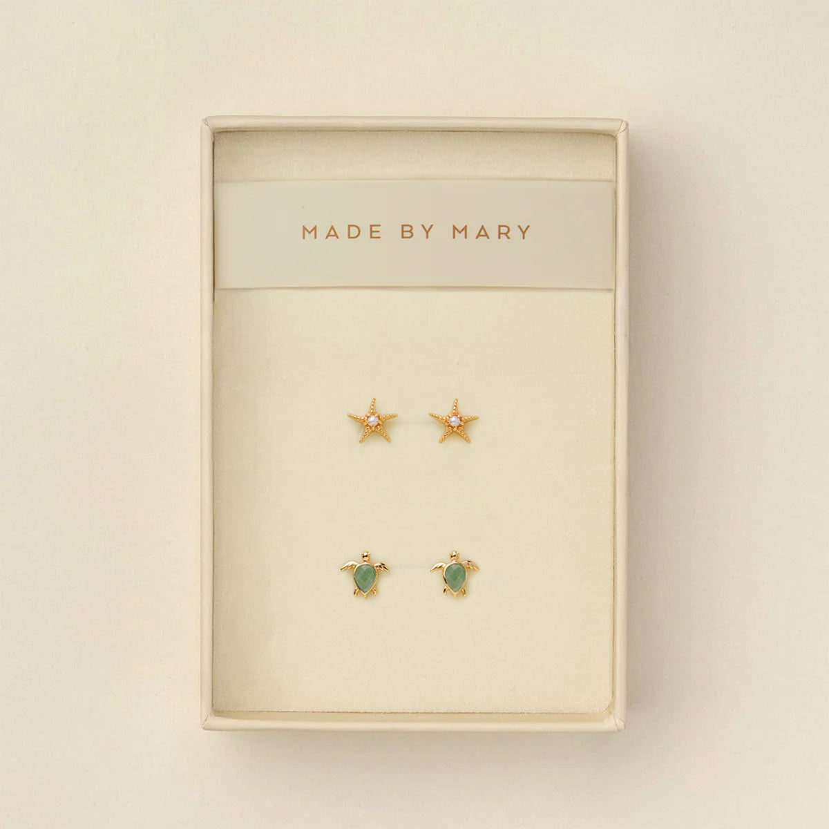 Set of gold stud earrings, one a starfish with a pearl in the center and the other a turtle with a green stone, in a 'Made by Mary' box on a beige background