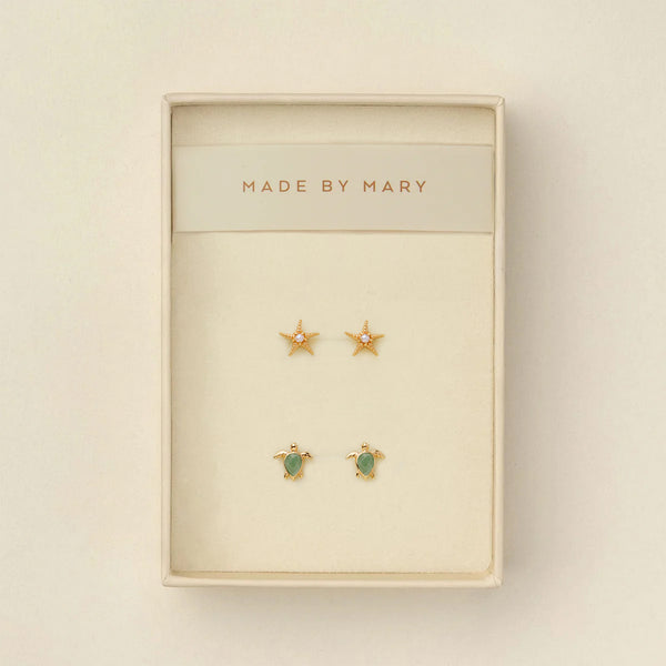 Set of gold stud earrings, one a starfish with a pearl in the center and the other a turtle with a green stone, in a 'Made by Mary' box on a beige background