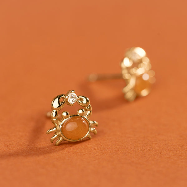 A close up of a gold crab stud earring, featuring an oval orange stone as the bodice and a small clear accent stone between it's claws. The background is a solid burnt orange color.