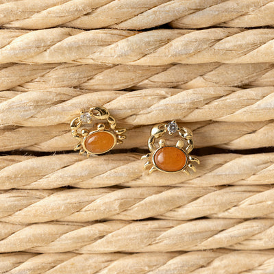 Gold crab-shaped stud earrings with orange stones on a woven straw background