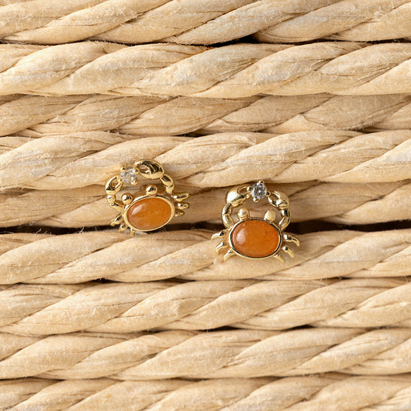 Gold crab-shaped stud earrings with orange stones on a woven straw background