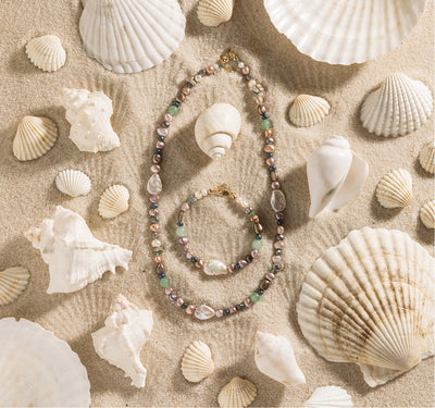 A multi-colored pearl necklace and bracelet set on sand surrounded by seashells