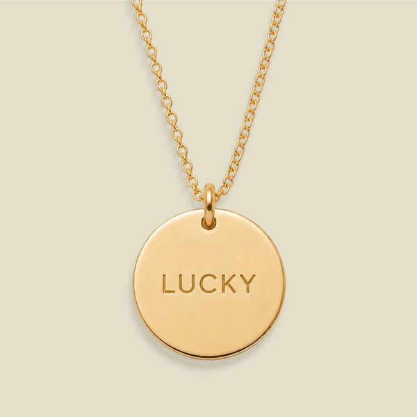 A gold chain necklace with a round gold disc pendant engraved with the word 'LUCKY' in capital letters, shown against a solid light beige background.