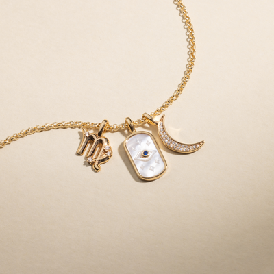 A gold chain necklace featuring a Virgo zodiac symbol charm, an oval charm with an eye design and white inlay, and a crescent moon charm covered in clear gemstones.